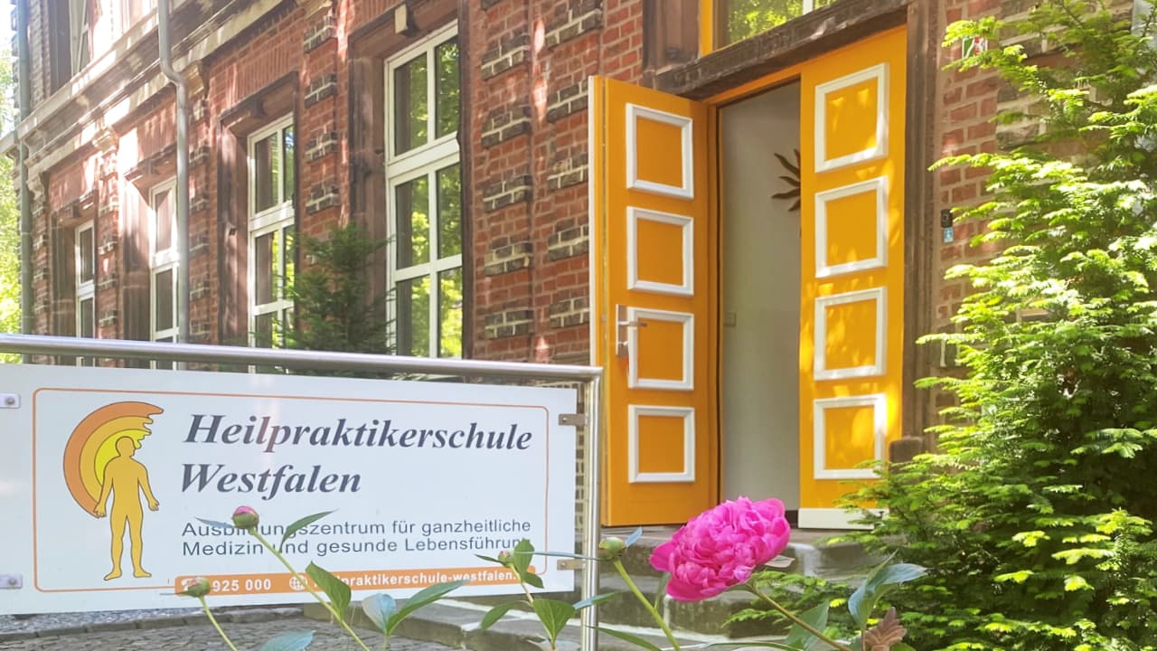 Eingang der Heilpraktikerschule Westfalen mit gelber Tür, Schulschild und Blumen im Vordergrund